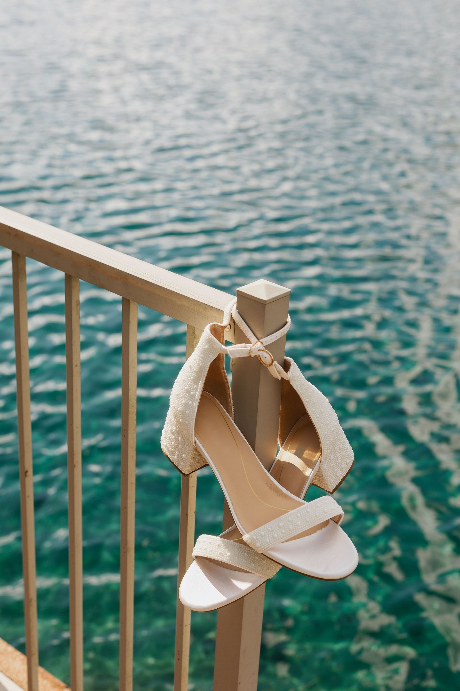 White bridal sandals displayed on railing overlooking turquoise water.