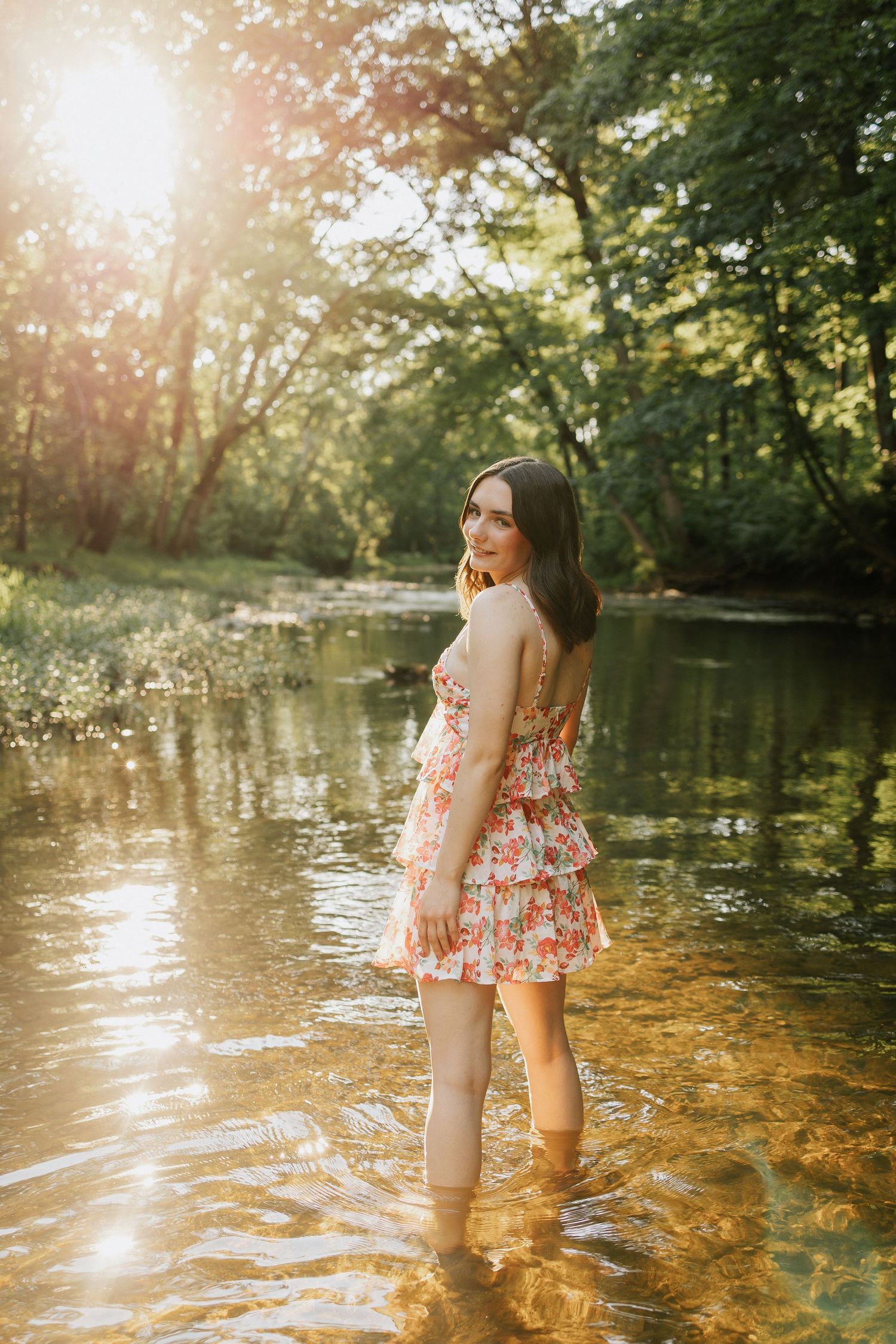 Young woman in floral dress standing in a sunlit forest creek, golden hour light filtering through green trees.