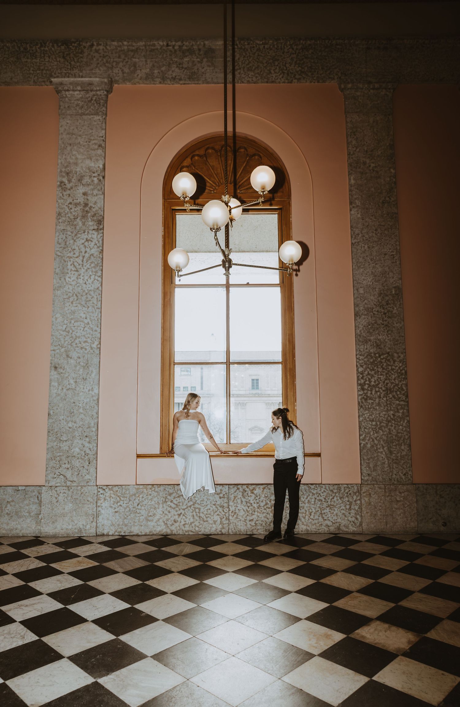 Bride and groom hold hands in elegant hall with checkered floors, arched window, and chandelier.