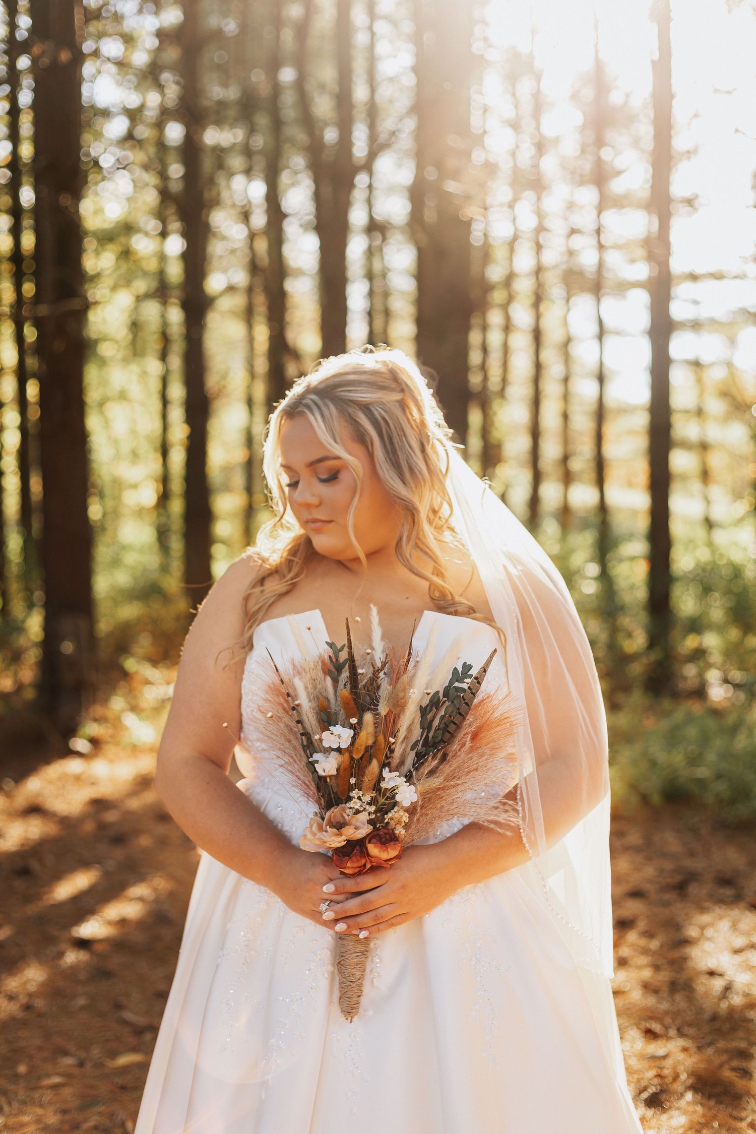 A bride in a white dress holds a rustic dried flower bouquet in a sunlit forest setting.