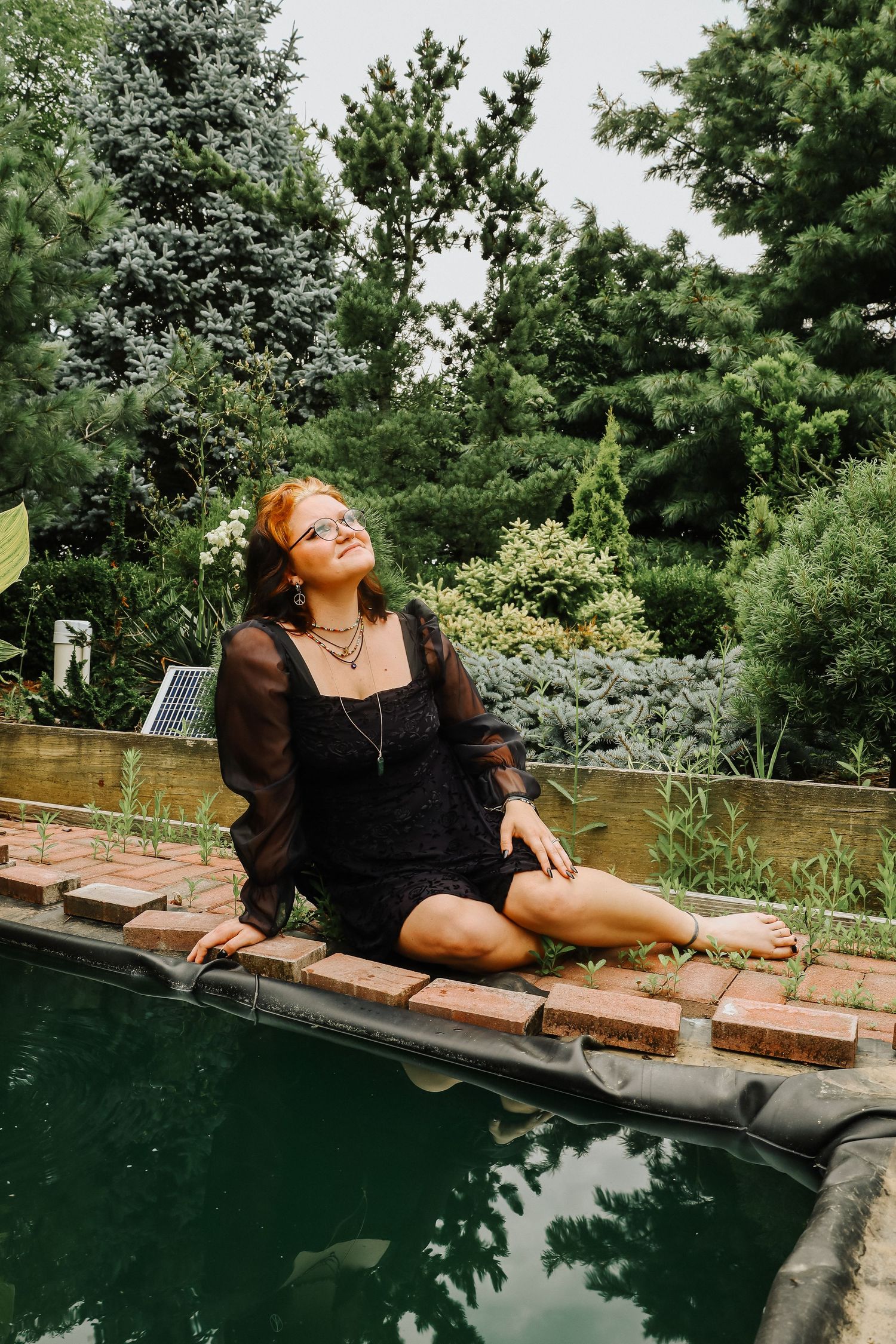 A person in black lounges along the edge of a dark pool surrounded by garden landscaping.