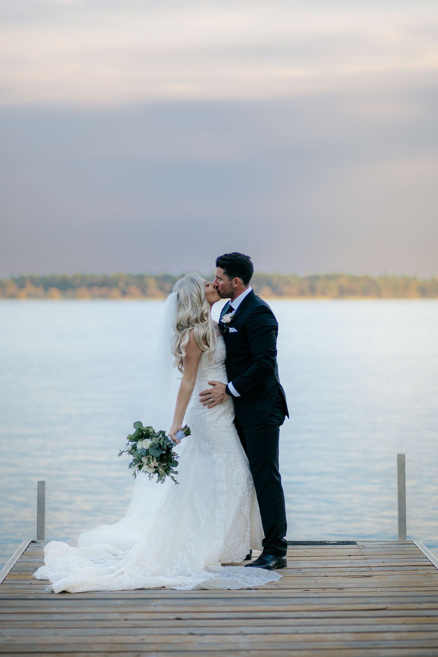 Newlyweds share intimate moment on wooden dock at sunset over serene lake.