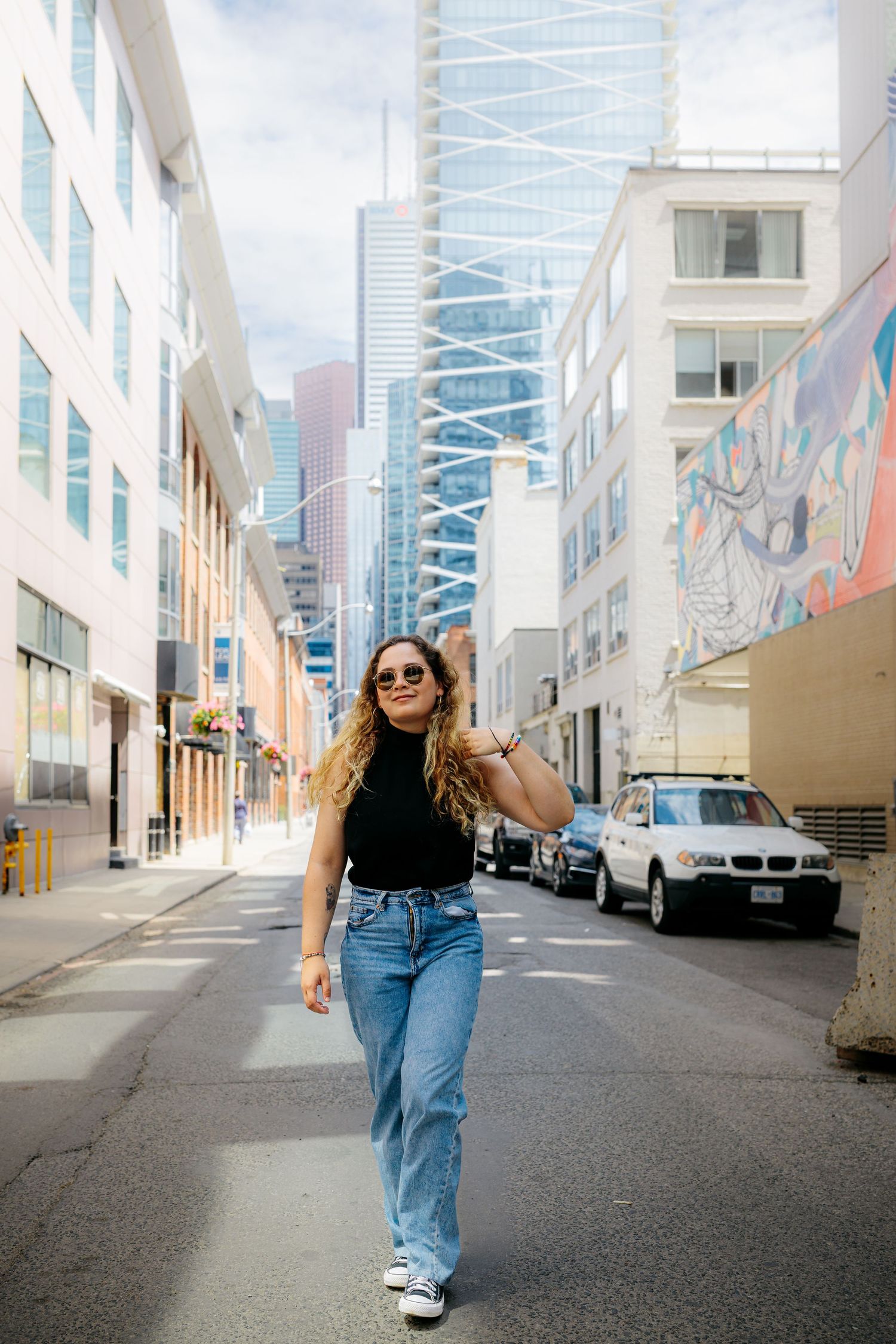 A casual street style photo in an urban alley with tall buildings and street art visible in the background.