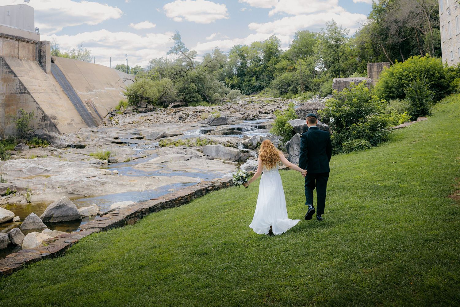 Couple walks along grassy riverbank near dam on their wedding day.