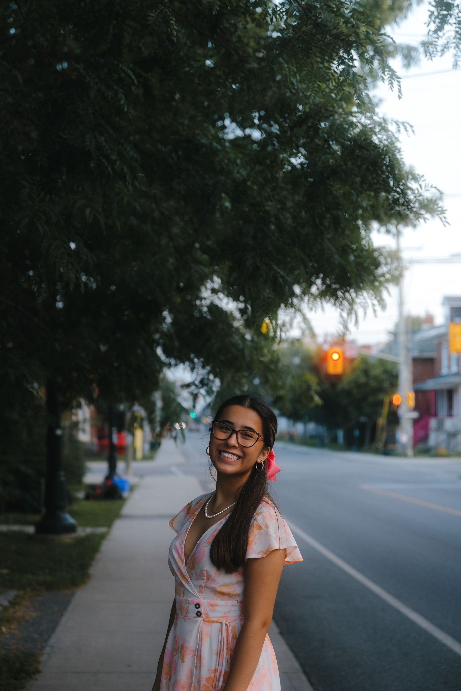 Street portrait of person in pink dress smiling on tree-lined sidewalk at dusk.