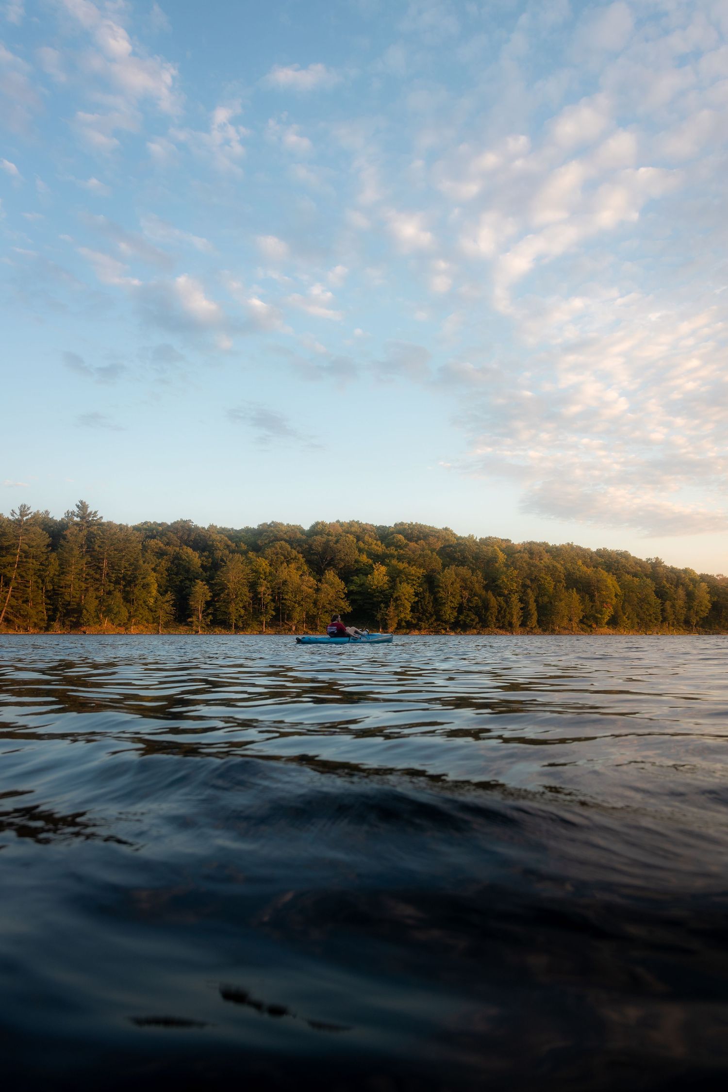 Calm lake water reflects forested shoreline under scattered clouds at sunset.