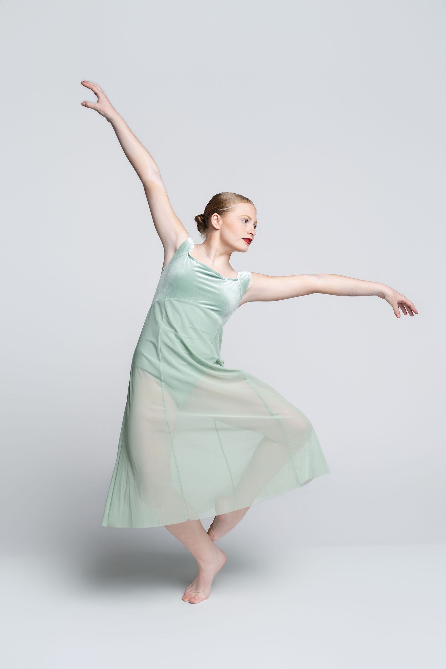 A graceful ballet dancer in a flowing mint green dress performs an elegant pose against a light gray background.