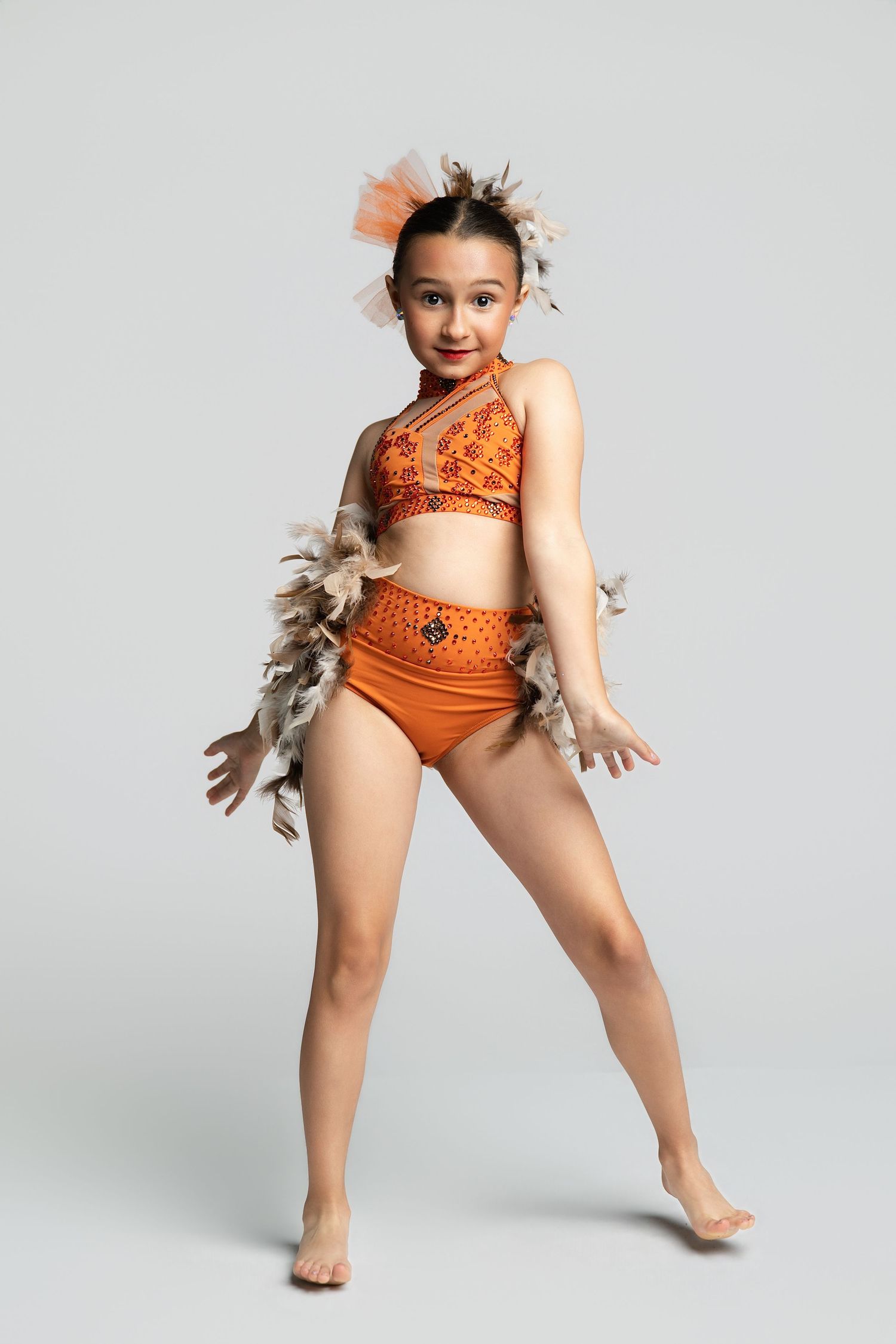 Young dancer in orange costume with feather accessories poses gracefully in a dance recital outfit.