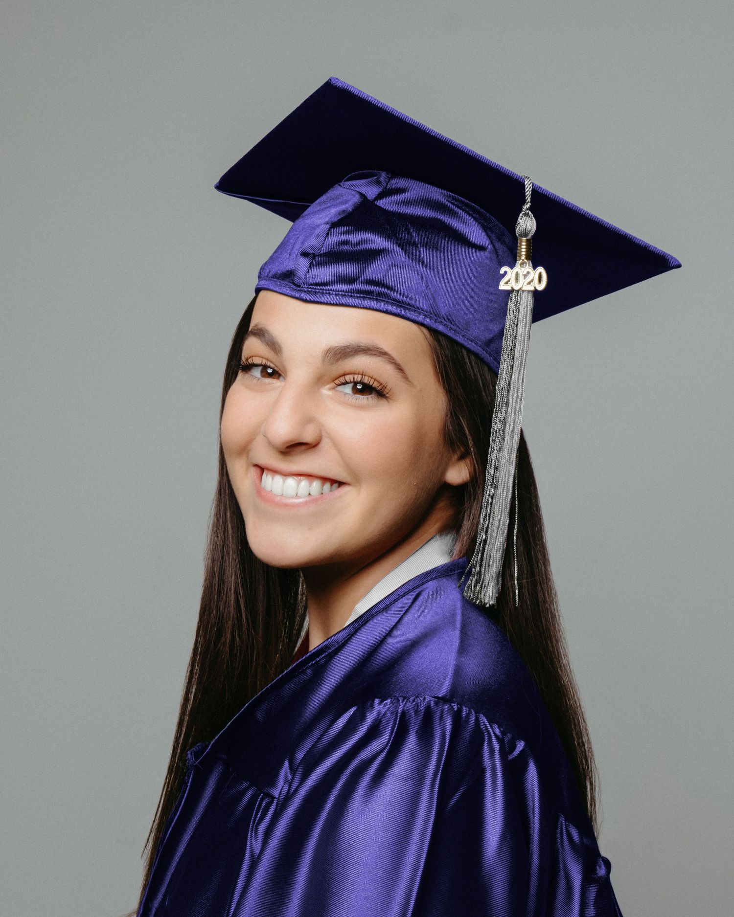 Graduate in purple cap and gown with 2020 tassel smiling against gray background.