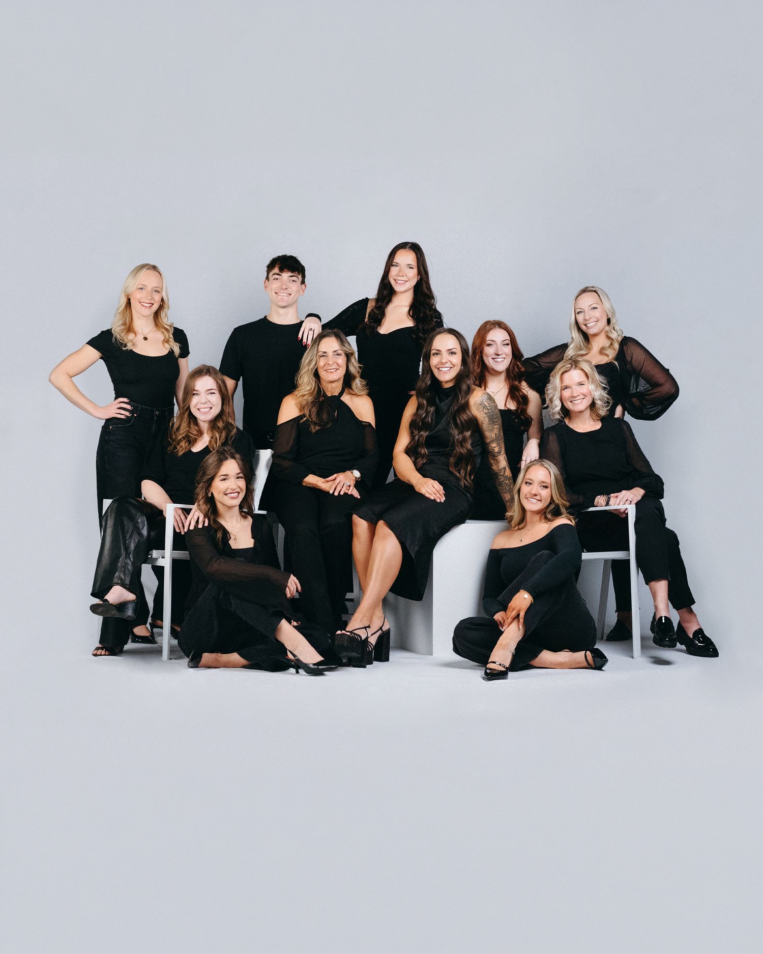 Group of eleven professionals posing together in coordinated black outfits against a light gray studio background.