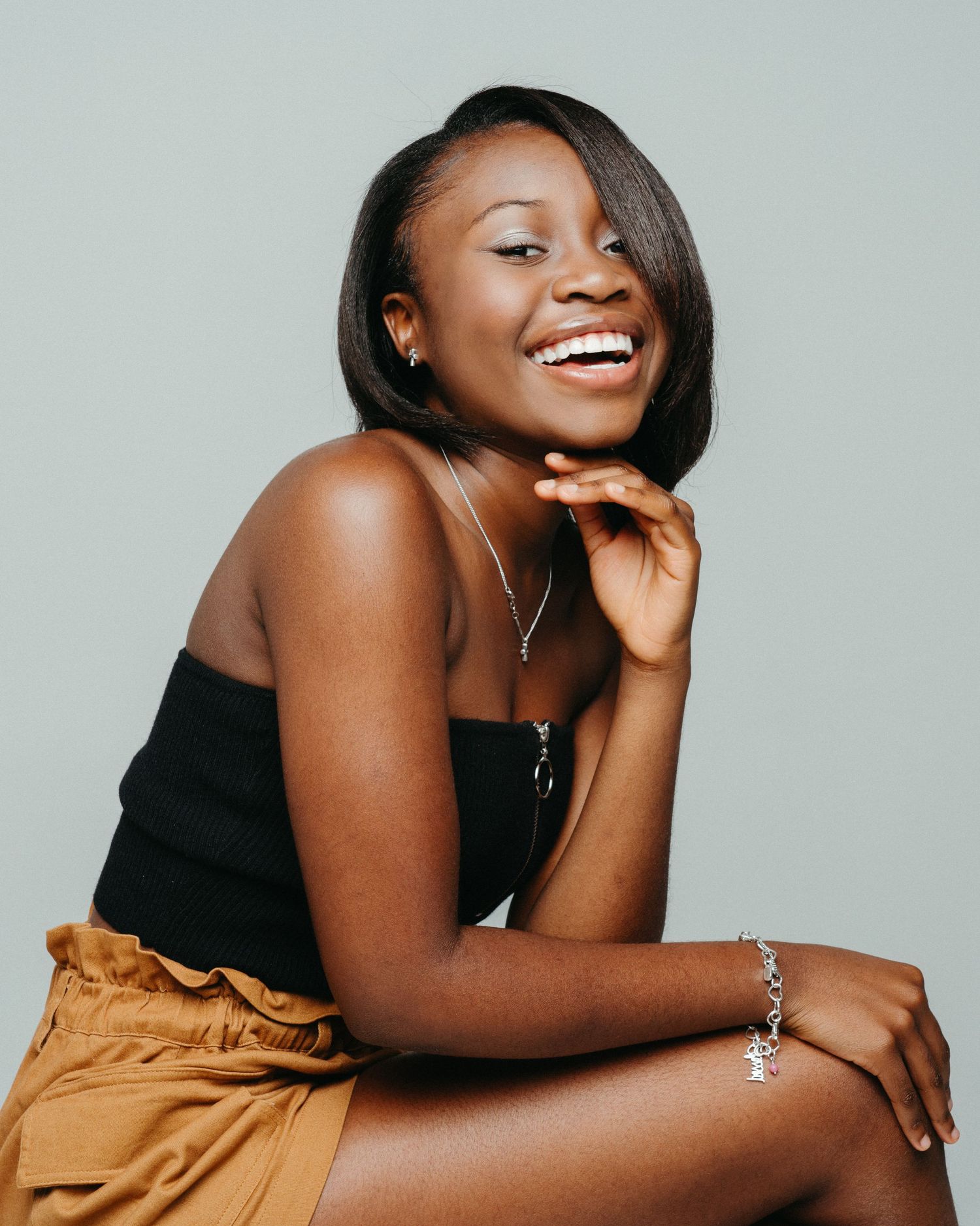 Stylish portrait of a person in black top and brown skirt smiling against gray background.