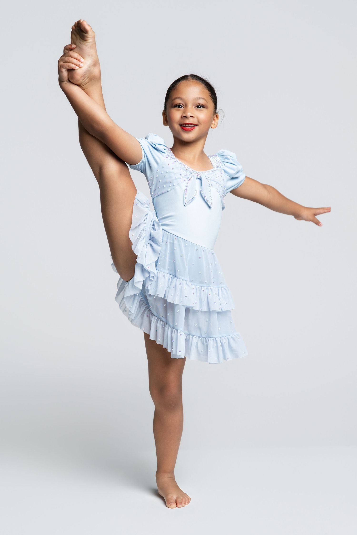 Young girl in light blue ruffled ballet dress performing a high leg extension while balancing on one foot.