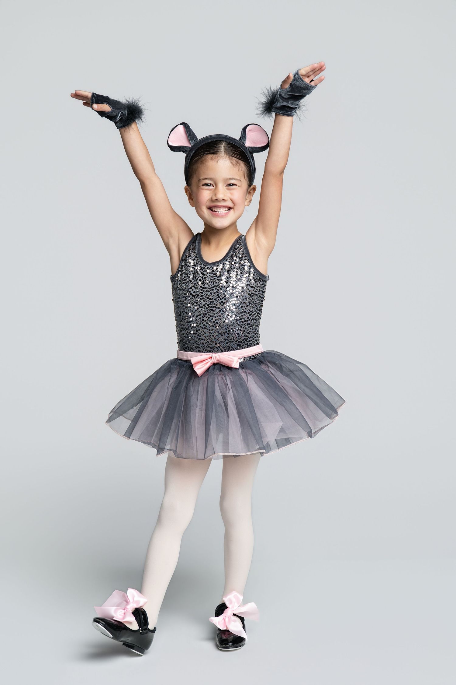 Young girl in sparkly black cat costume with tutu, ears, and pink bows poses ballet stance on gray background.