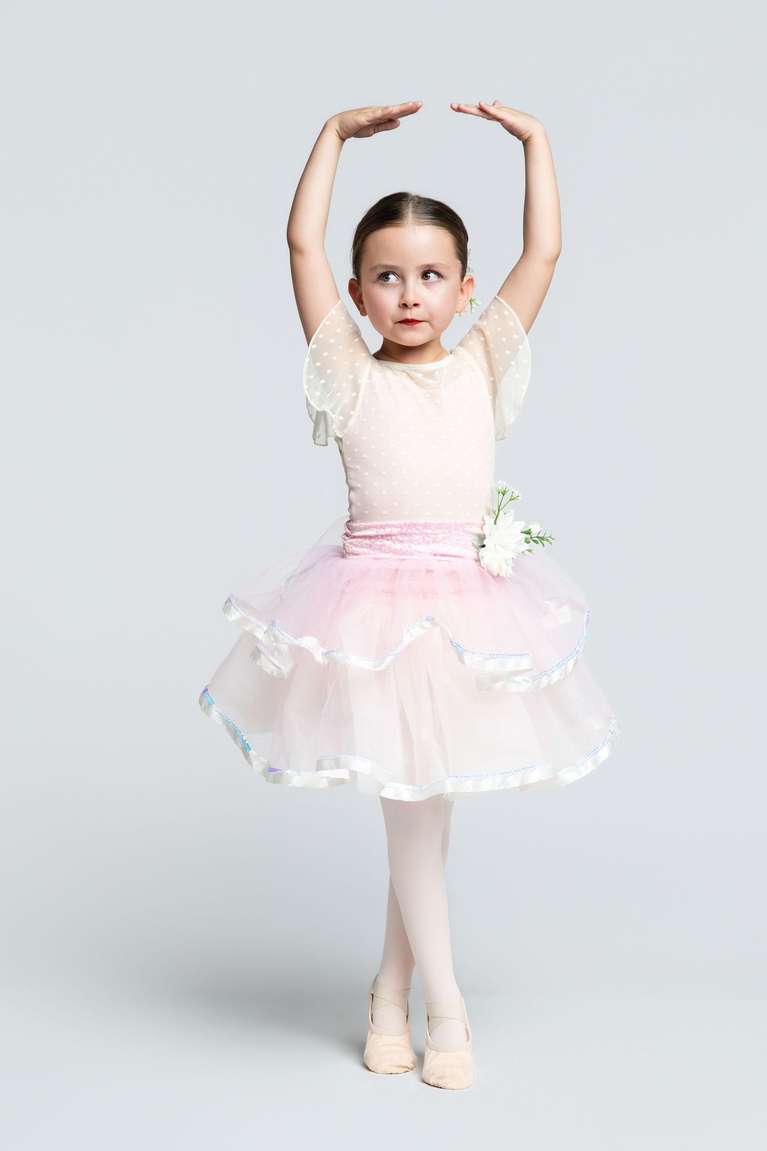 Young girl in pink tutu and ballet shoes poses gracefully with arms raised overhead in classic ballet stance.