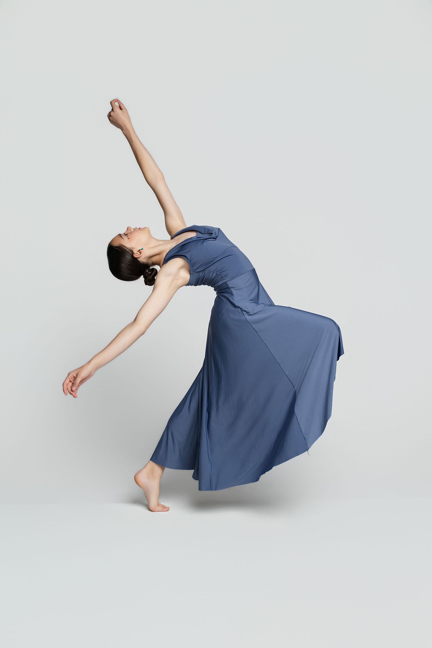 A barefoot dancer in a flowing blue dress performs an elegant backbend with one arm raised against a white background.