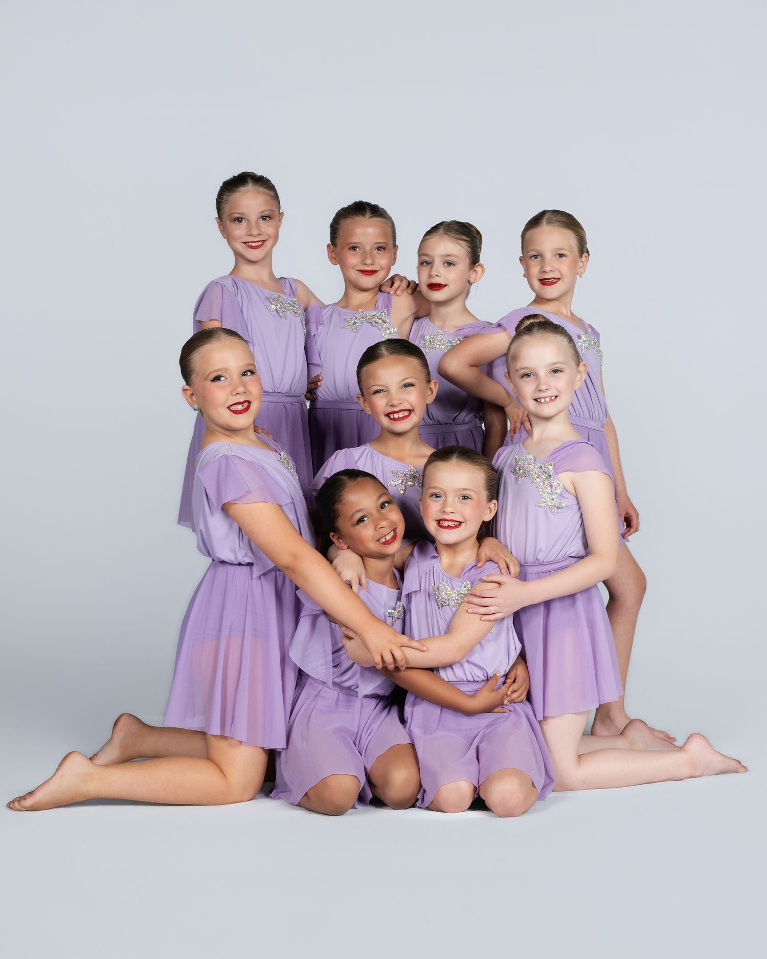 Eight young girls in matching lavender dance costumes pose together for a group dance recital portrait photo.