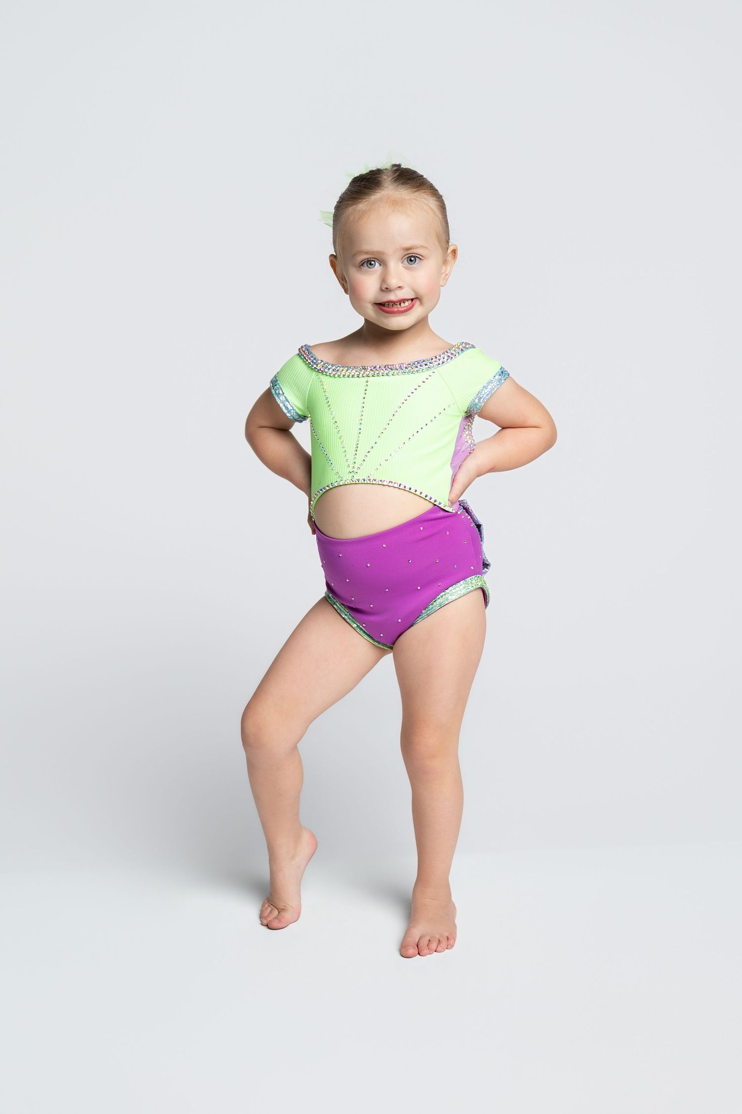 Young girl in green crop top and purple shorts posing confidently with hands on hips against white background.