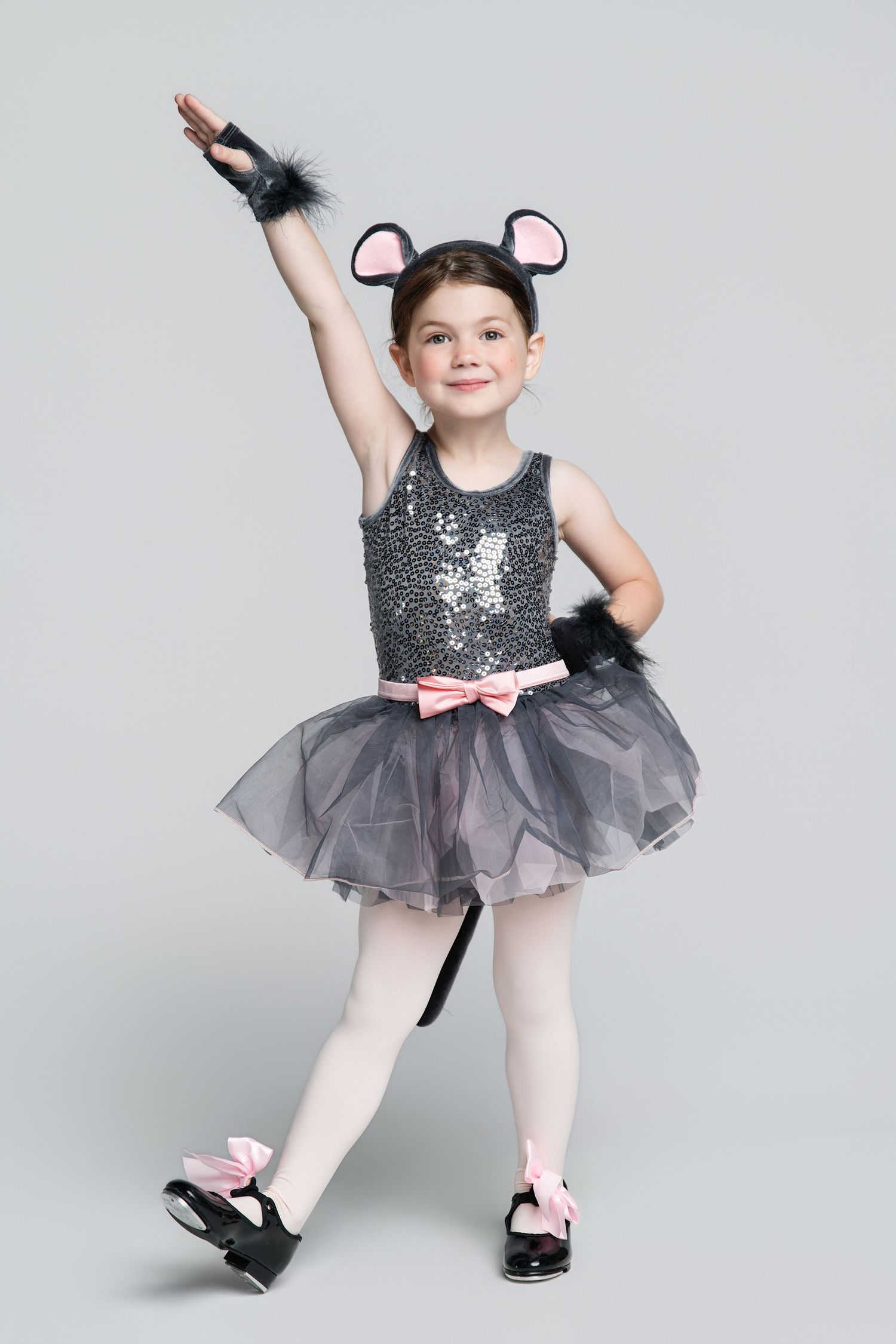 Young girl in sequined cat costume with tutu, mouse ears, and tail poses in ballet stance on gray background.