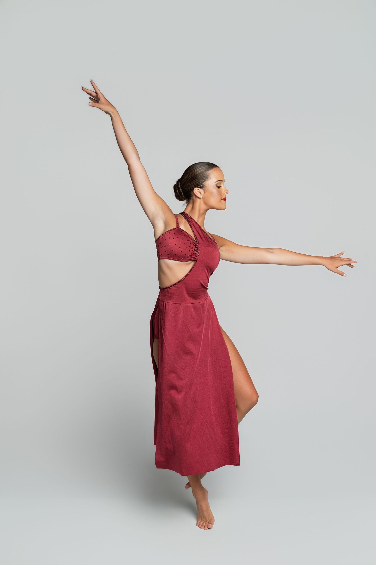Graceful dancer in a burgundy dress performs an elegant pose with arms extended on a gray background.