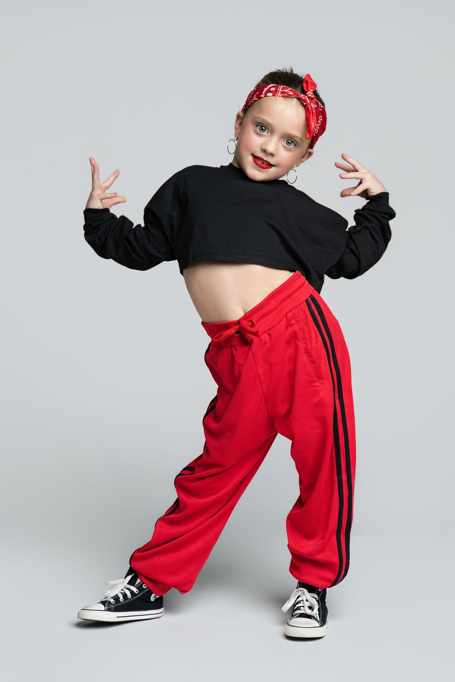 Young girl in red baggy pants and black crop top striking a hip-hop dance pose on gray background.