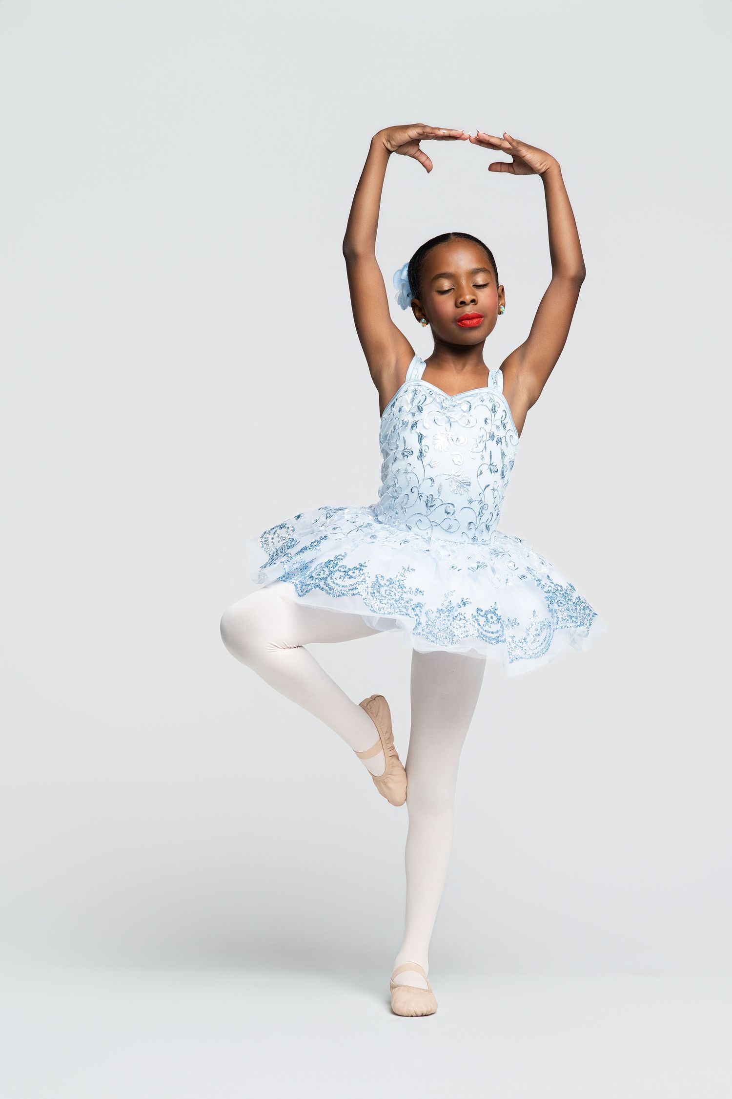 Young Black ballerina in blue floral tutu performs elegant pose on one leg with arms raised overhead.