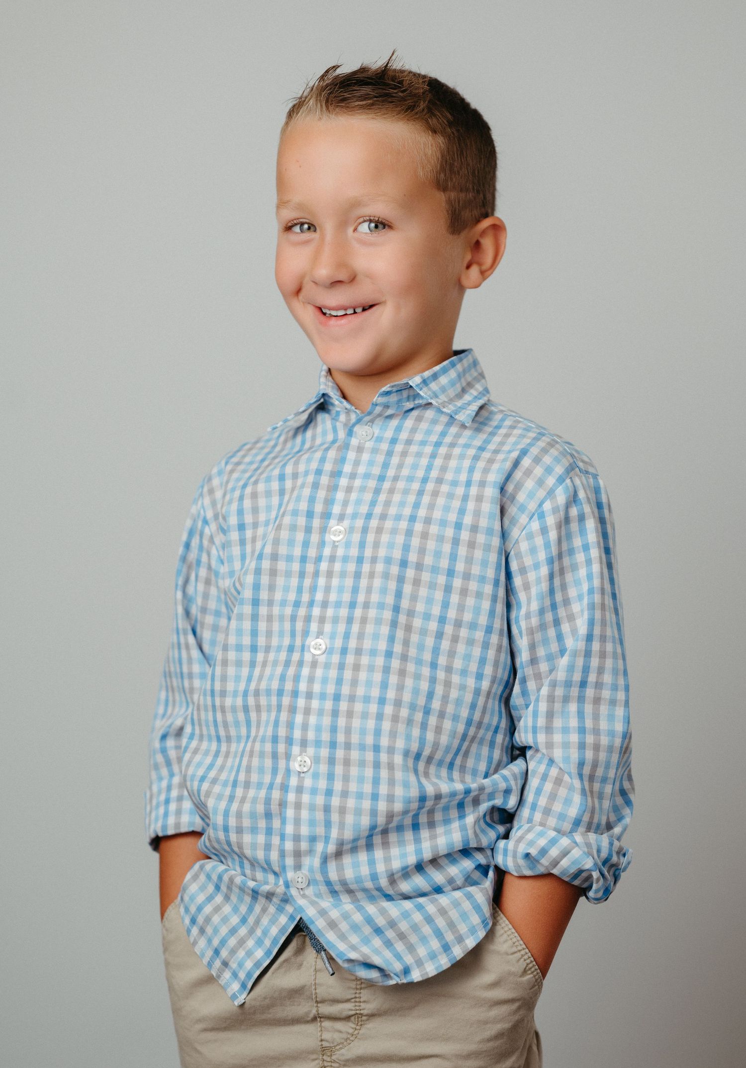 Young student wearing a light blue checkered button-down shirt smiling against a gray background.