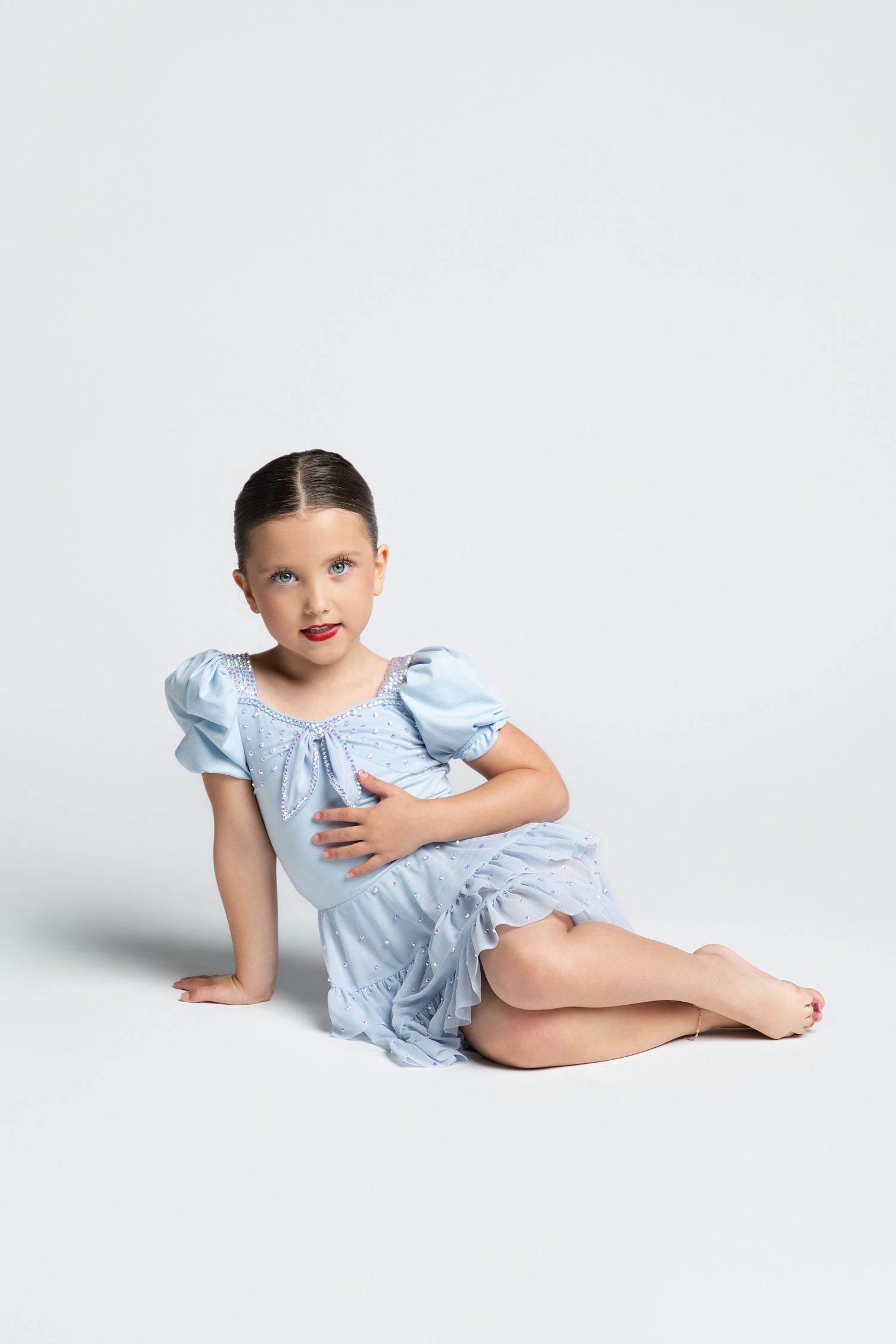 Young girl in a light blue puff-sleeve dress sits gracefully on white floor, looking at camera.