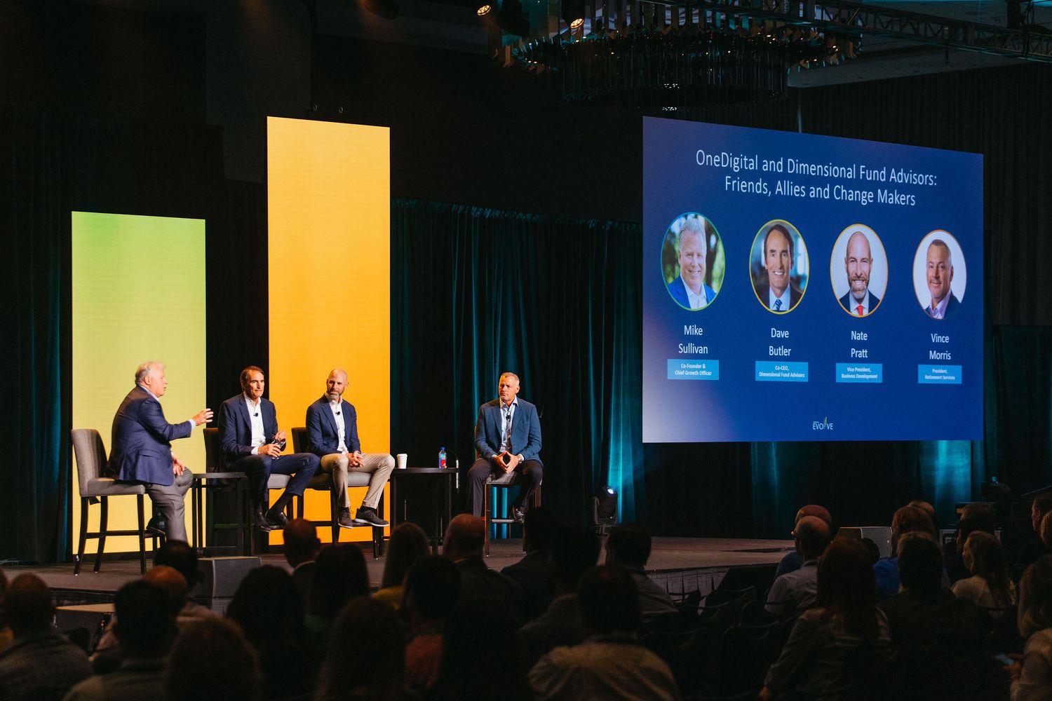 Conference stage setup with blue lighting and multiple speakers seated at a panel discussion.