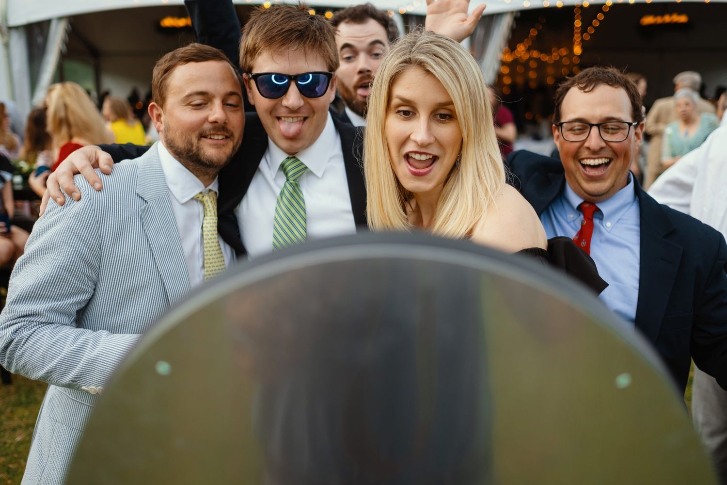 Group of friends posing and smiling at an outdoor wedding reception under a white tent.