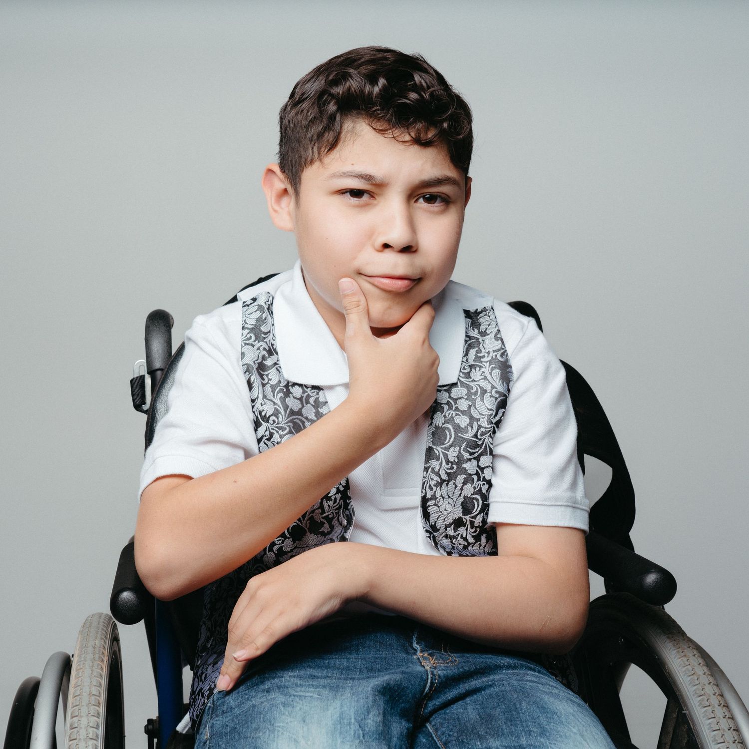 Young person in wheelchair wearing vest and white shirt with thoughtful expression.