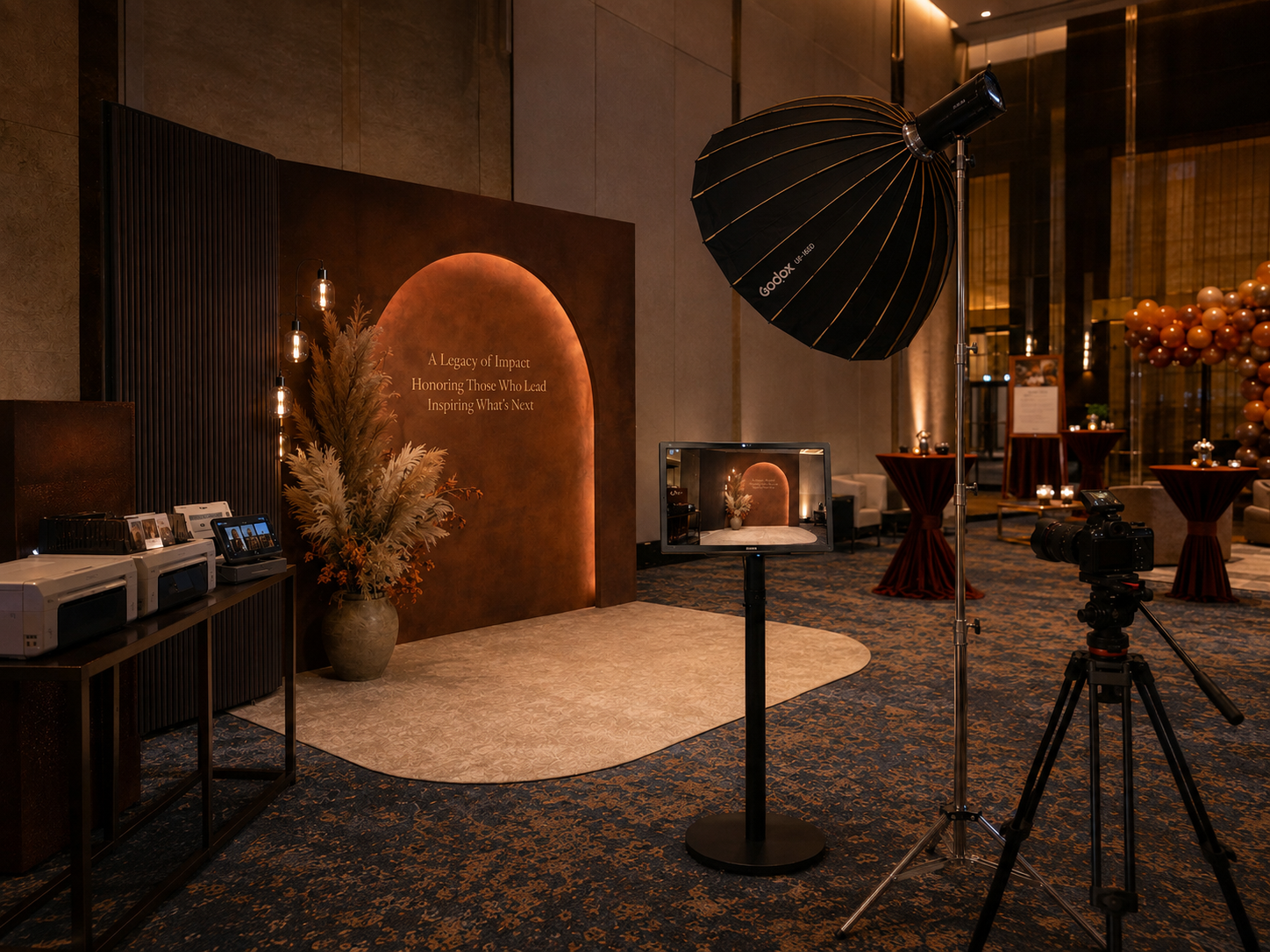 Elegant event space with warm lighting, decorative arch backdrop, photography equipment, and balloon decor in amber tones.