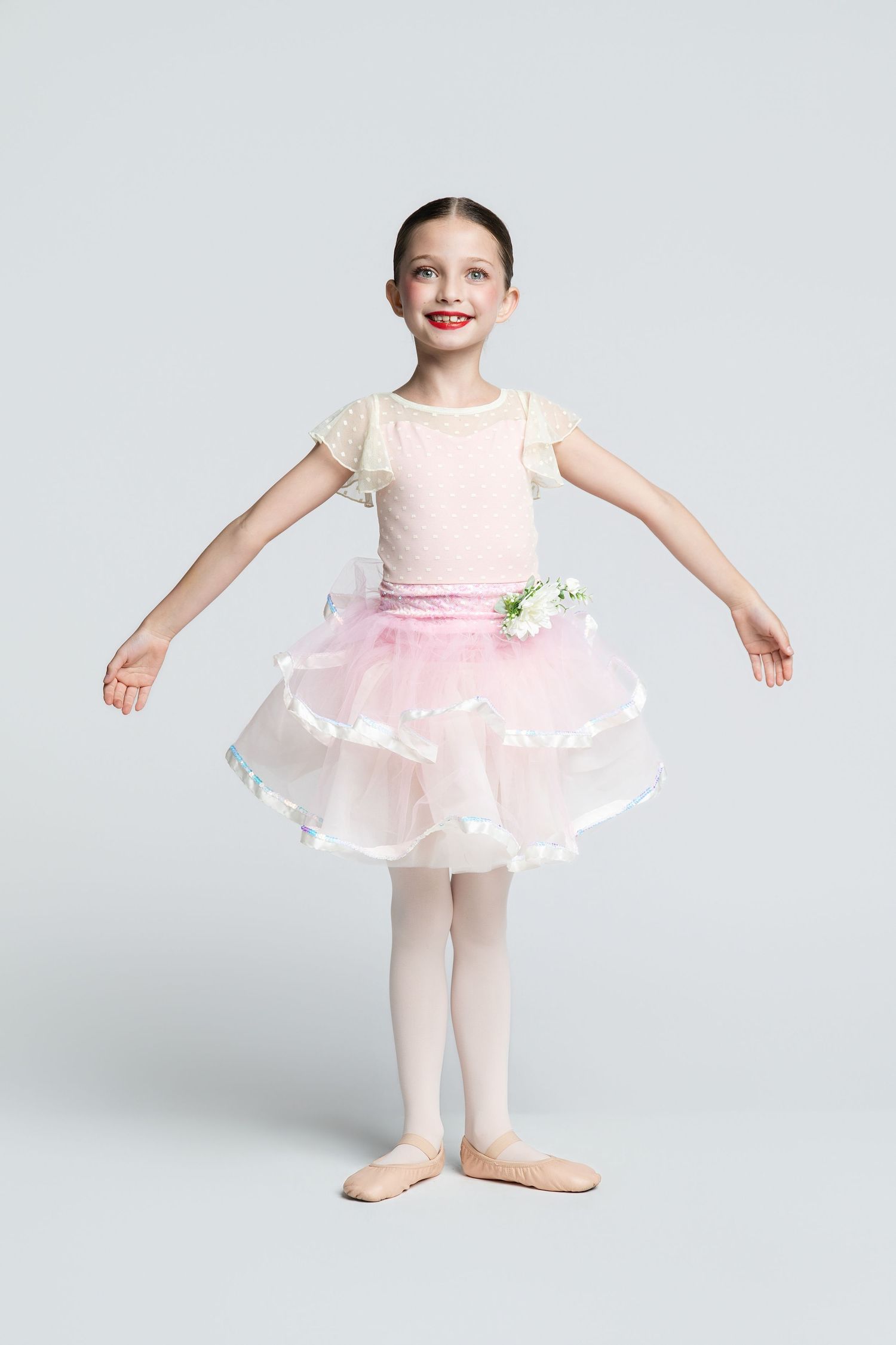 Young girl in pink tutu and ballet slippers poses gracefully in a dance studio, smiling brightly.