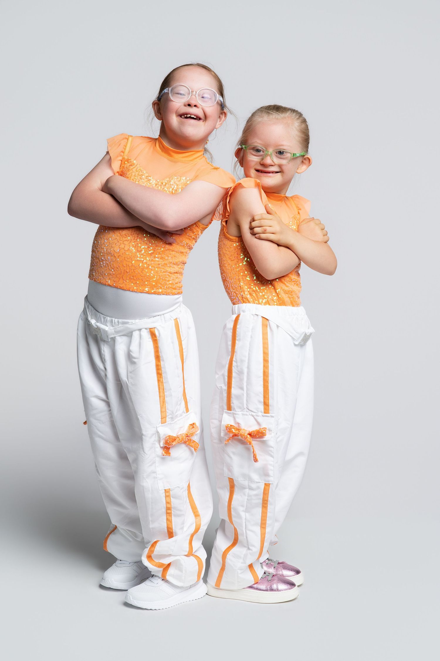 Two young girls with Down syndrome wearing matching orange tops and white pants with orange trim, smiling and posing together.