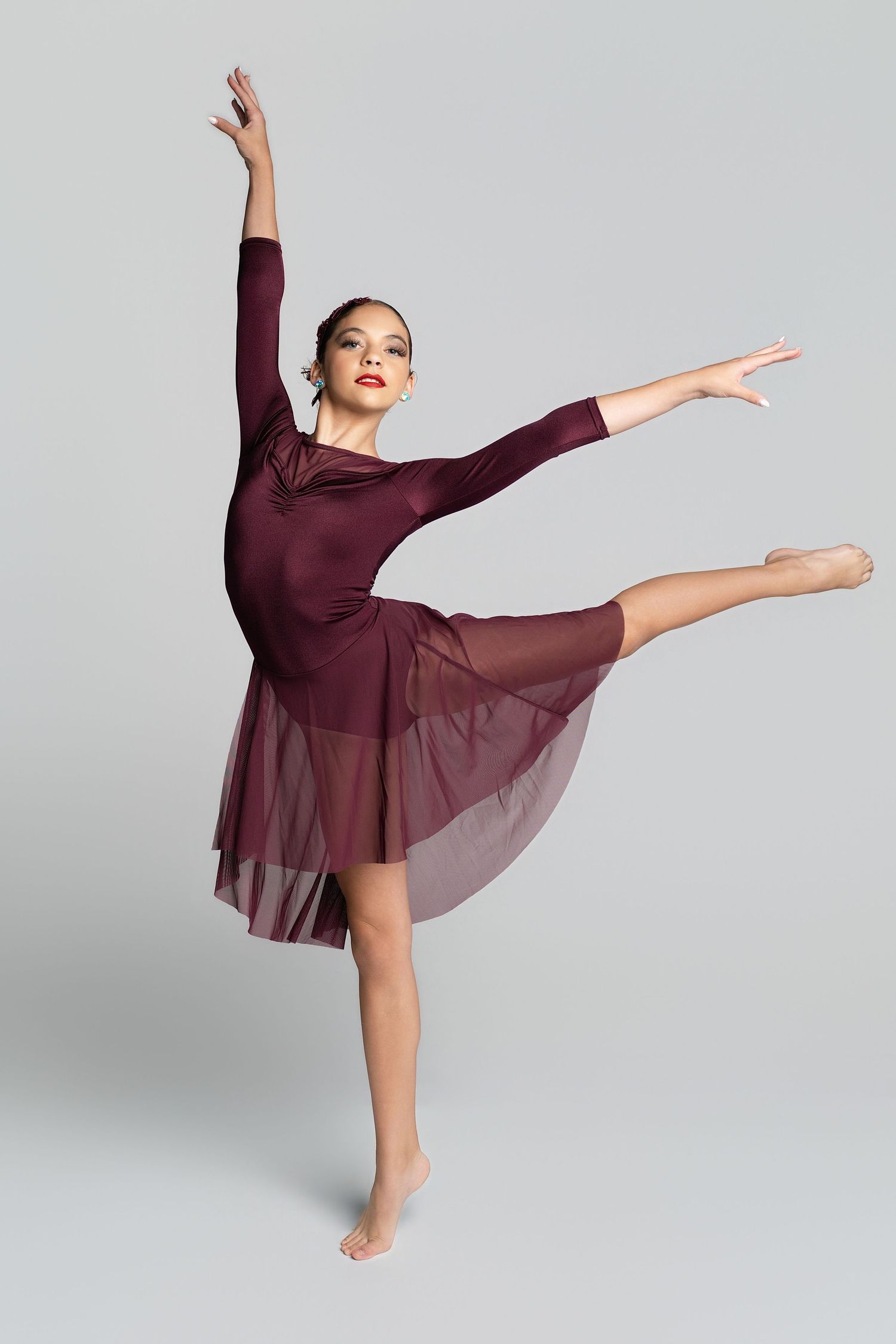 A ballet dancer in a burgundy dress performs an elegant arabesque pose against a gray background.