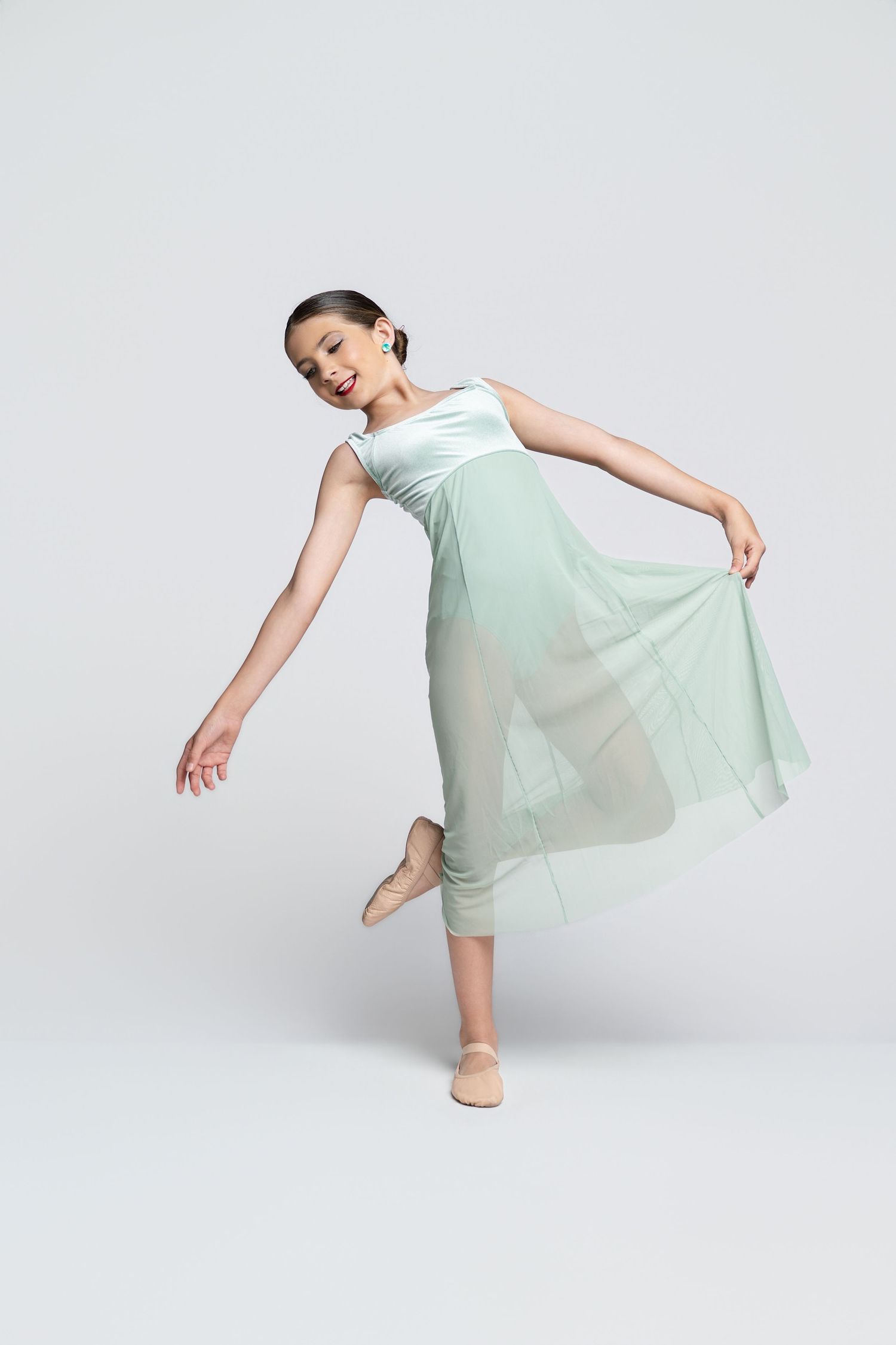 Young ballerina in mint green chiffon dress performing a graceful ballet pose on one foot against white background.