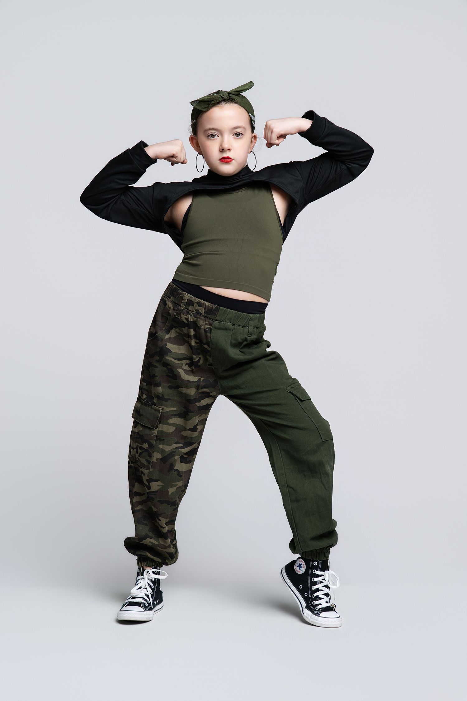 Young girl in hip-hop dance pose wearing camo cargo pants, crop top, black sneakers, and green headband.