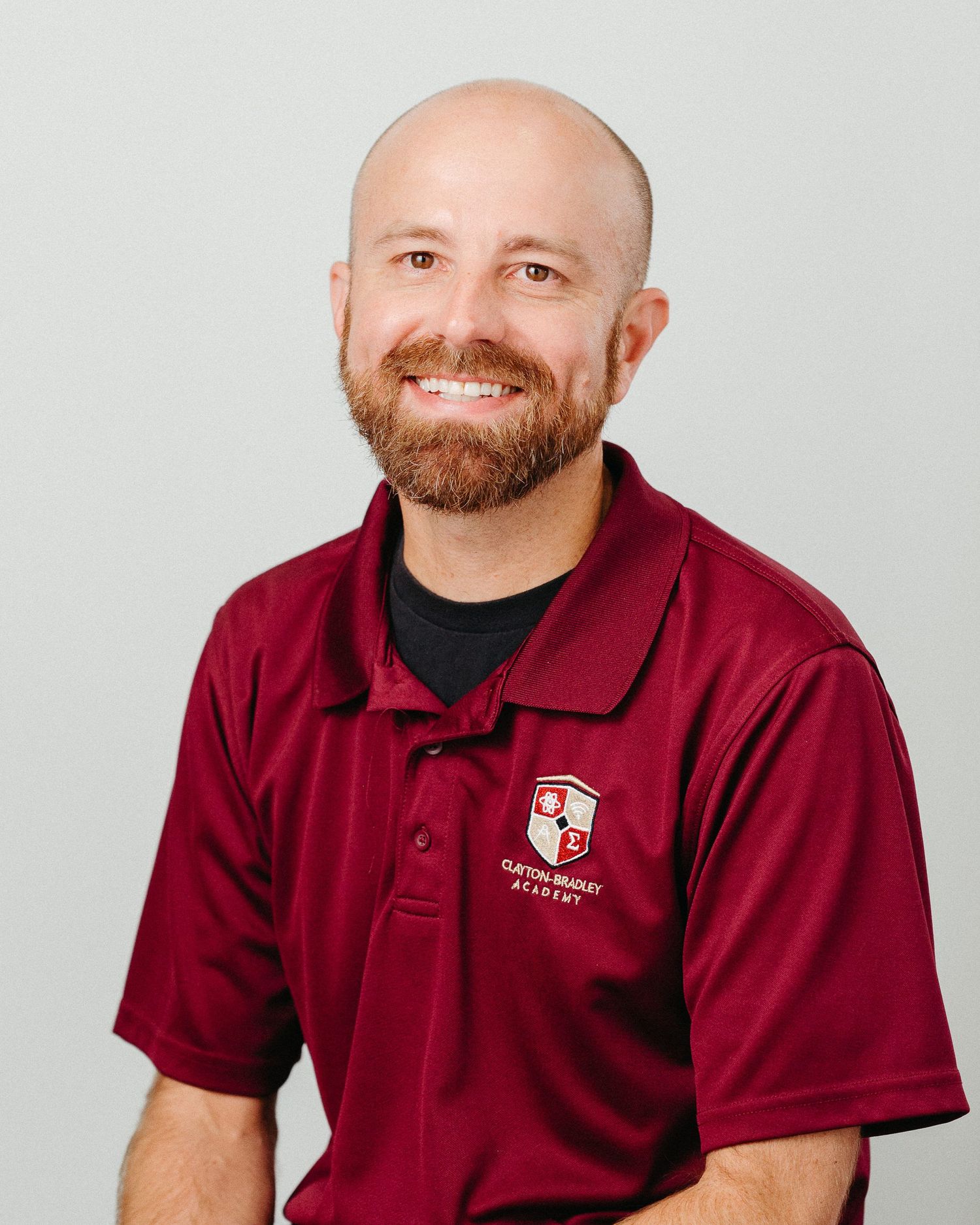 Staff member in burgundy polo shirt with school logo smiles warmly against light gray background.