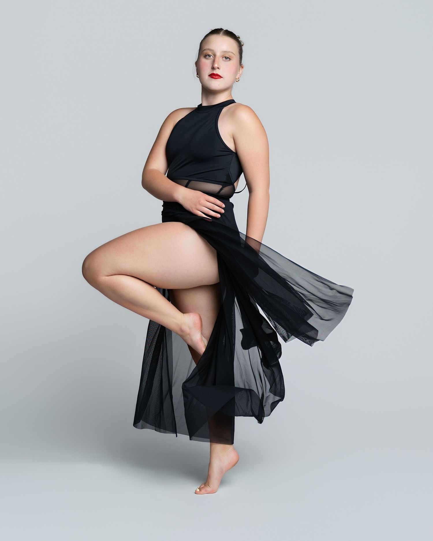 Dancer in black halter top and flowing sheer skirt poses gracefully on one leg against gray background.