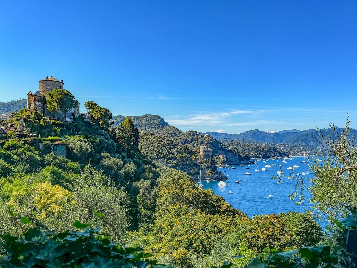Scenic hilltop chapel overlooking Portofino bay with lush greenery and sailboats in the blue harbor.