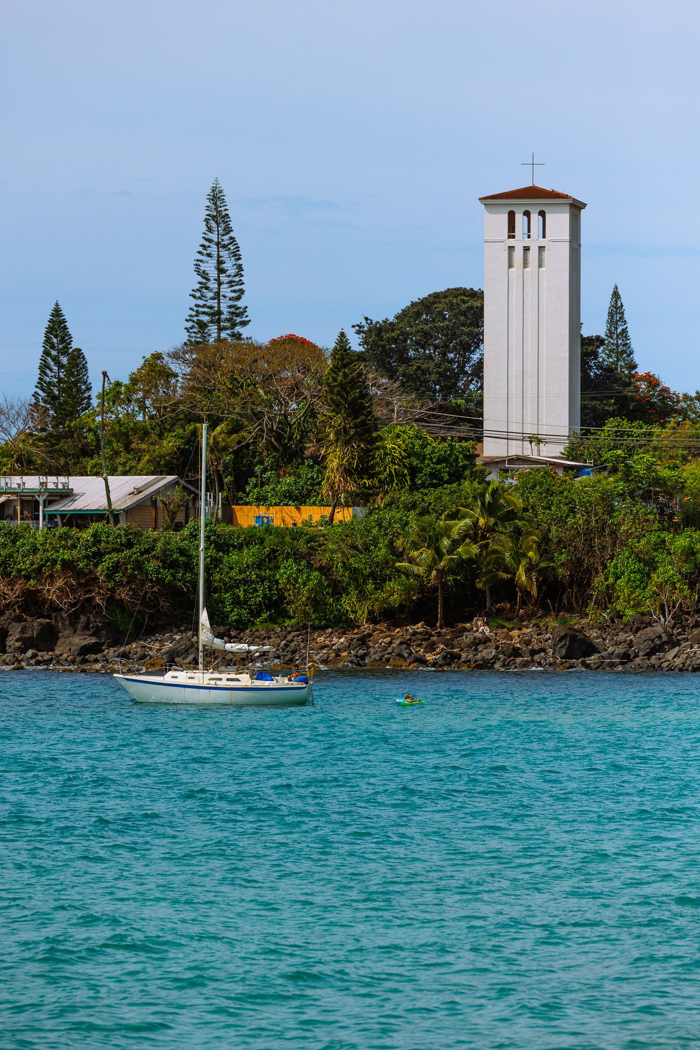 Sailboat anchored in turquoise waters near a lush tropical shoreline with a white church bell tower among pine trees.