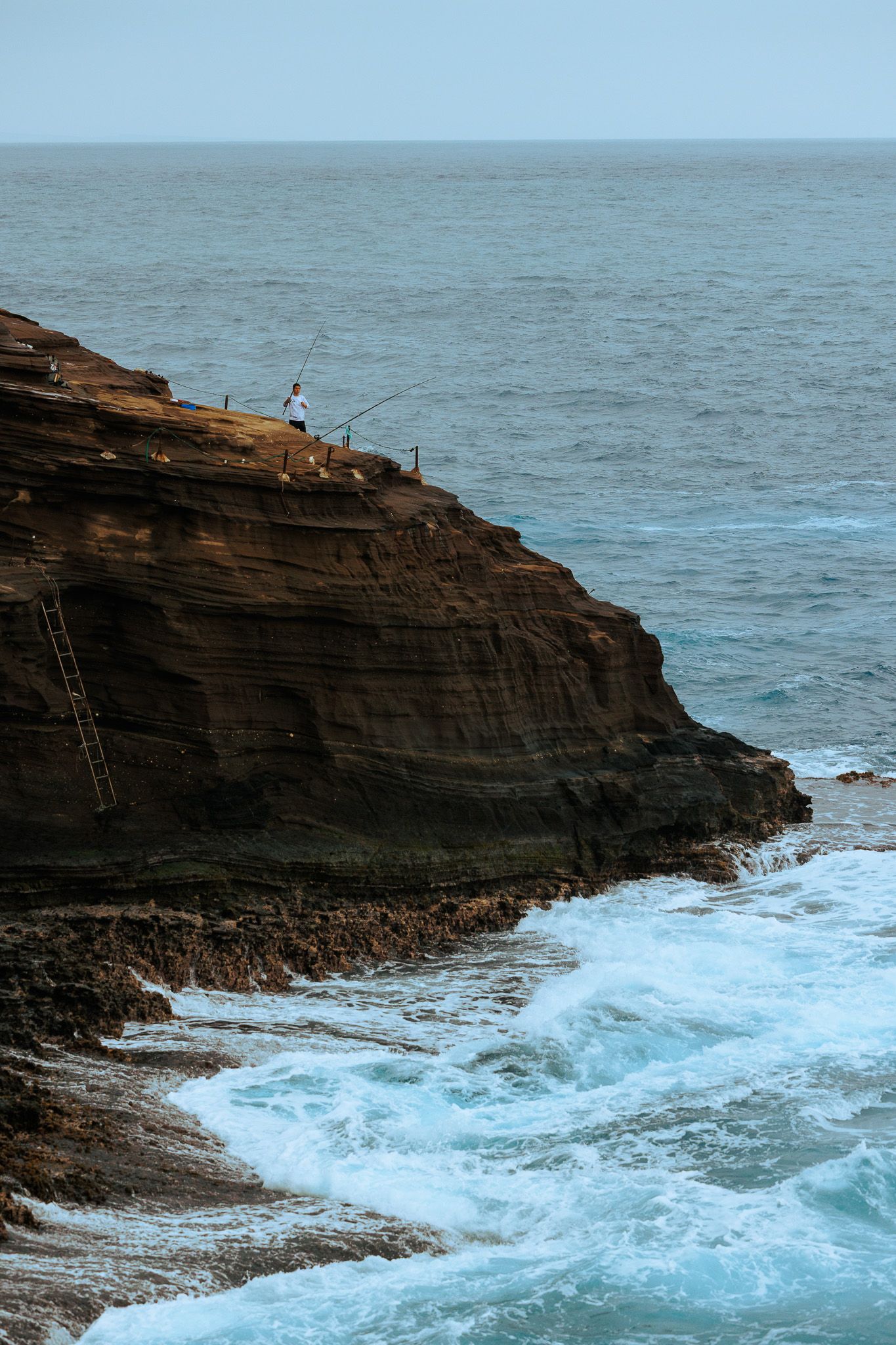 Person fishing on a rugged coastal cliff edge above crashing ocean waves, with a metal ladder on the rocky seaside.