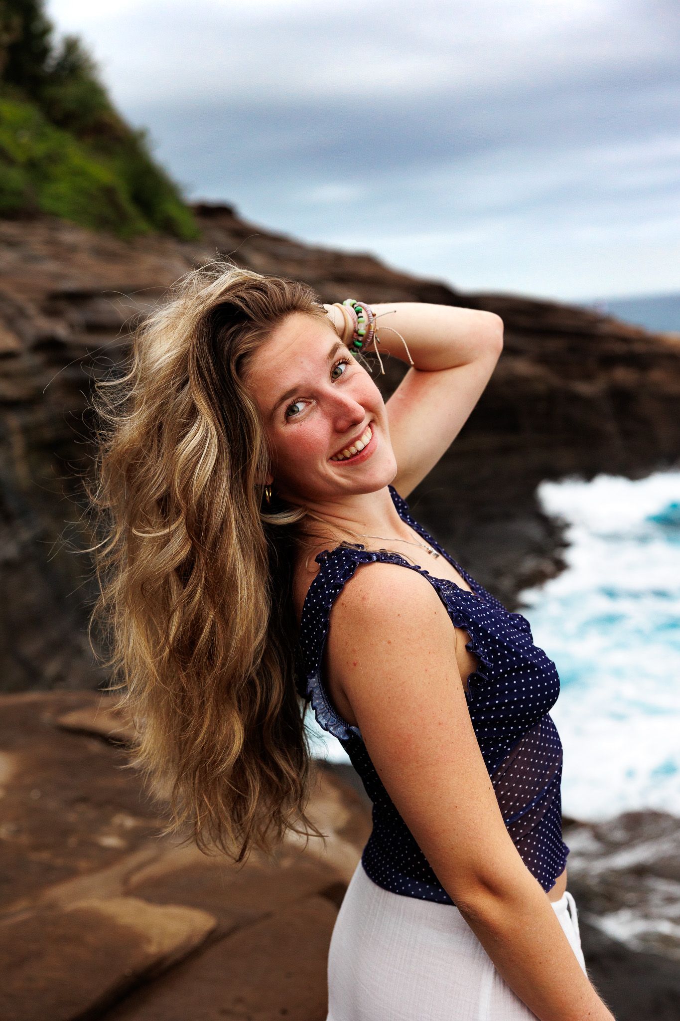 Happy woman with flowing long hair smiling at rocky seaside cliff with ocean waves behind her.