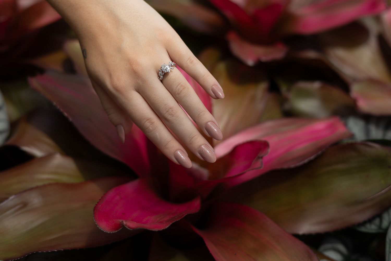Close-up of a diamond engagement ring on a woman's hand resting on vibrant red tropical leaves.