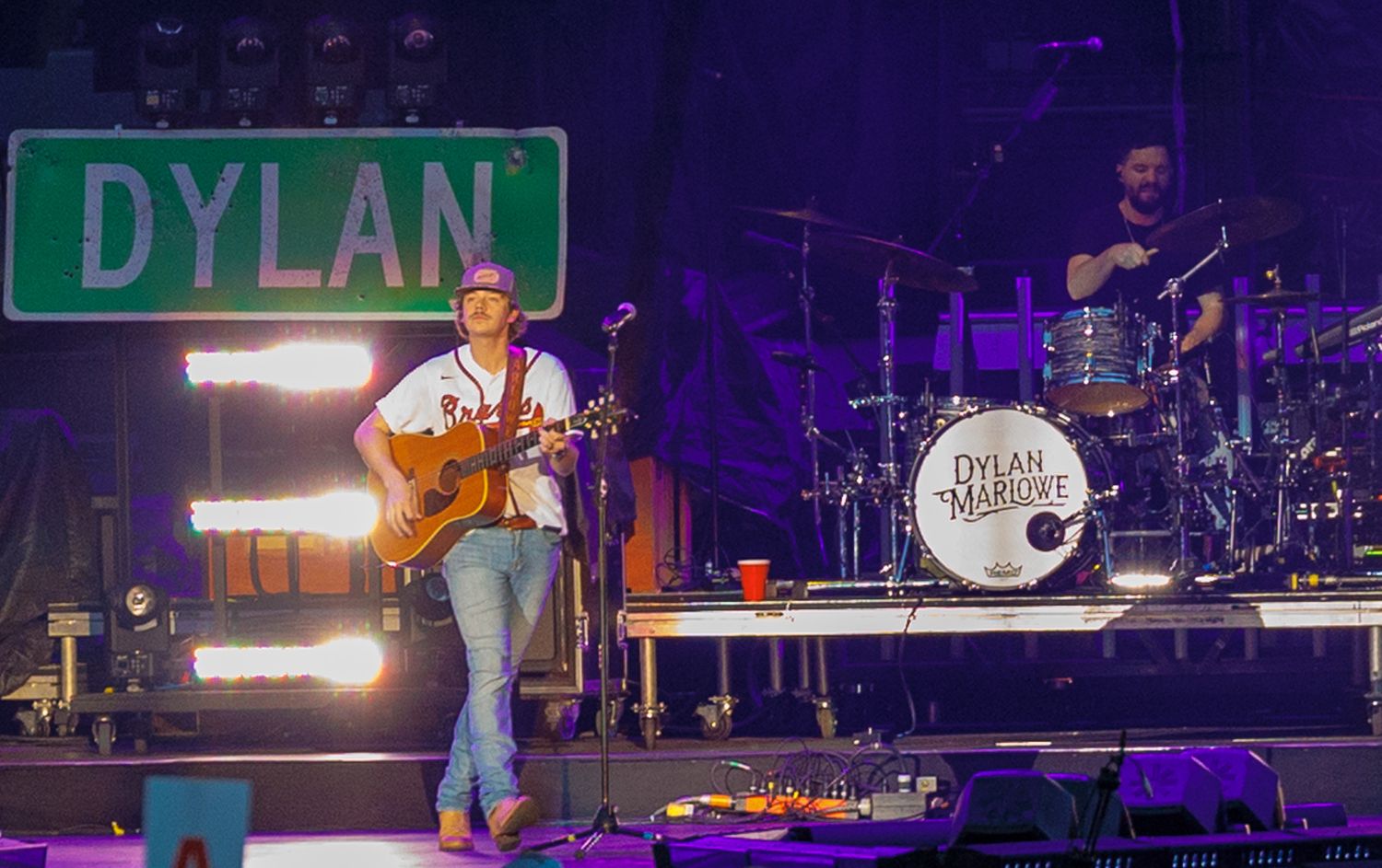 Dylan Marluke performing live on stage with guitar and drummer behind a green Dylan street sign.