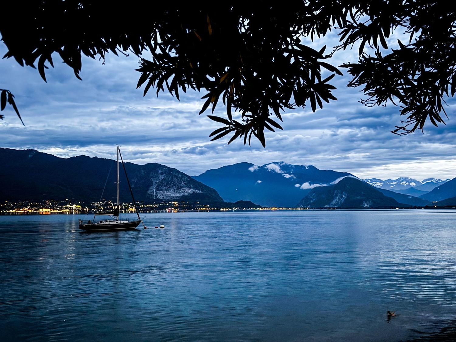 Sailboat floats on calm lake at dusk with snow-capped mountains and city lights behind.