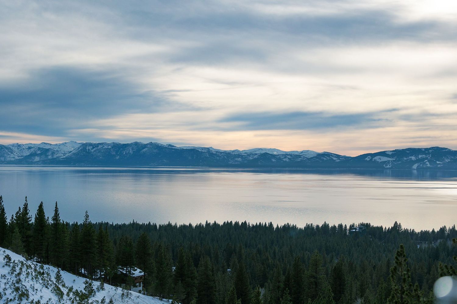 Panoramic winter sunset view of Lake Tahoe with snow-covered pine trees and mountain backdrop.
