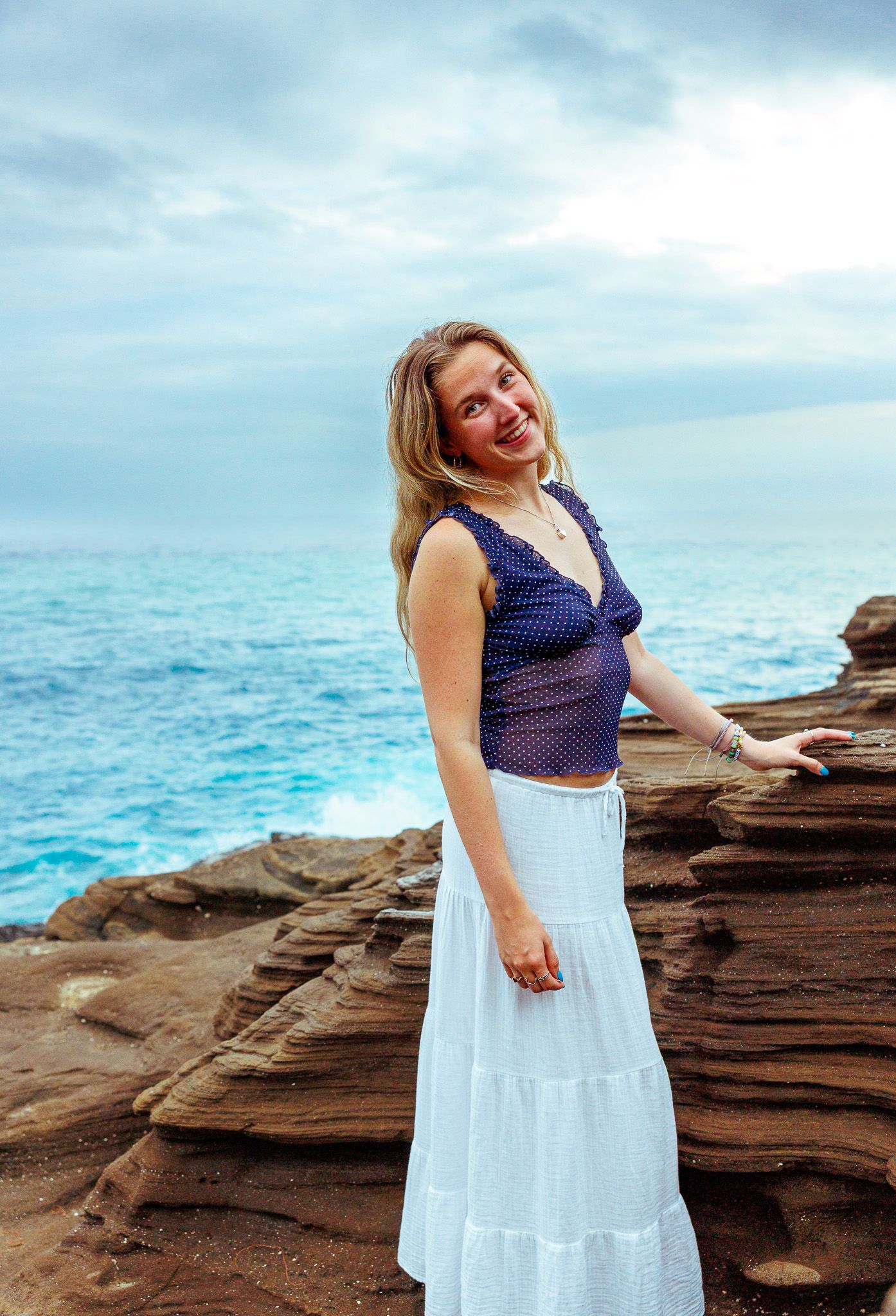 Smiling blonde woman in navy polka dot top and white maxi skirt standing on layered red rock sea cliffs.