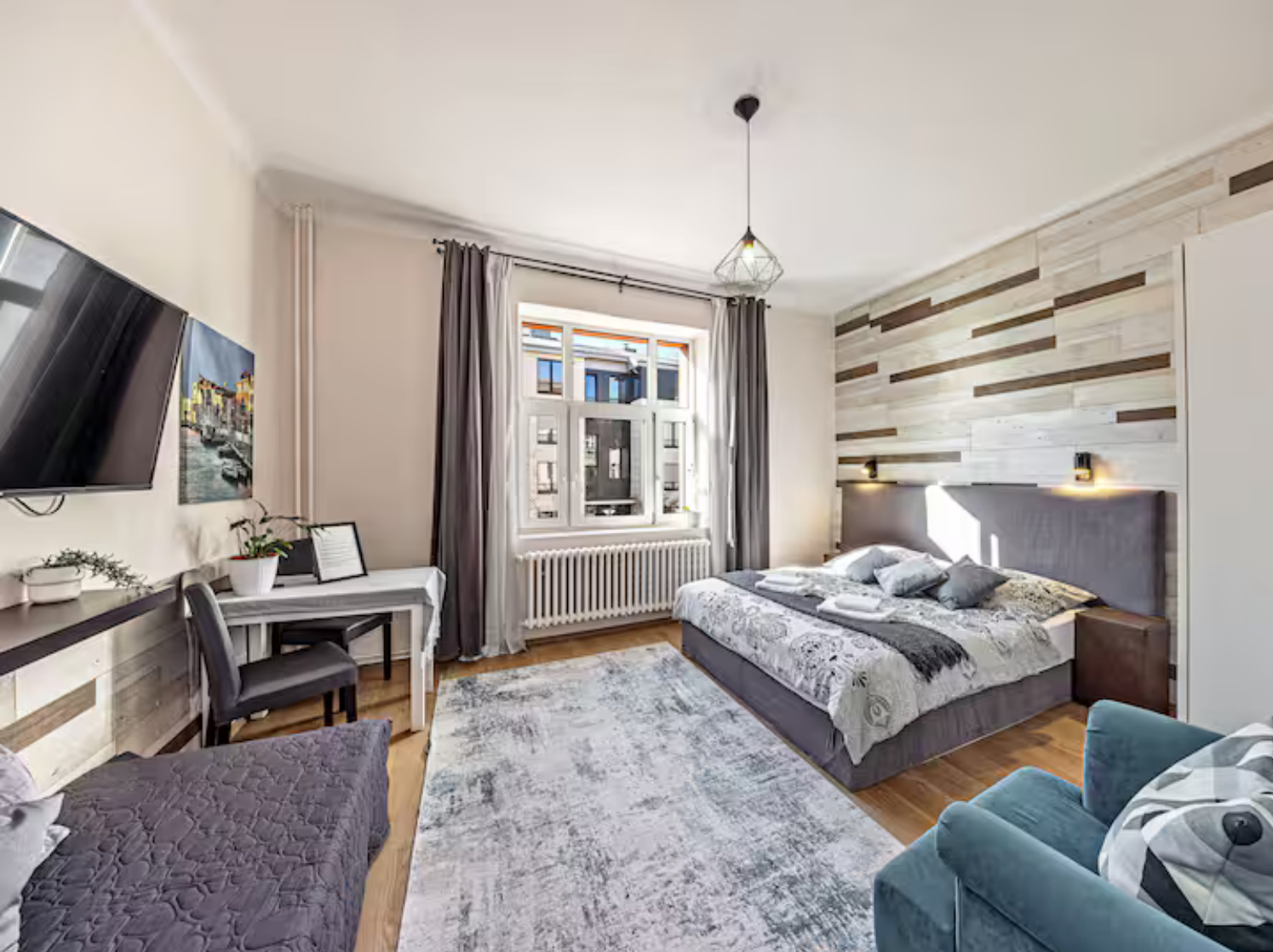 Modern hotel studio bedroom with accent wood wall, queen bed, desk, TV, and blue sofa on hardwood floors.