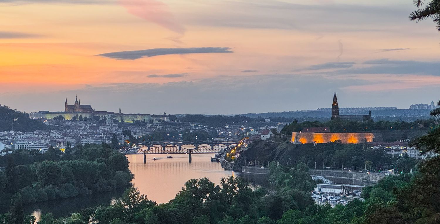 Panoramic sunset view of Prague skyline with the Vltava River and historic castle glowing at dusk.