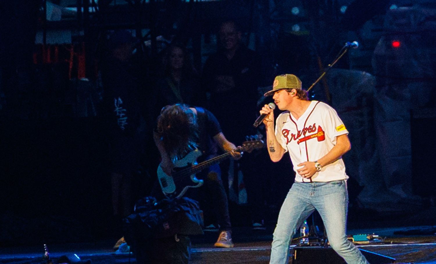 Singer in Braves jersey and hat performs with microphone while guitarist plays behind him on stage.